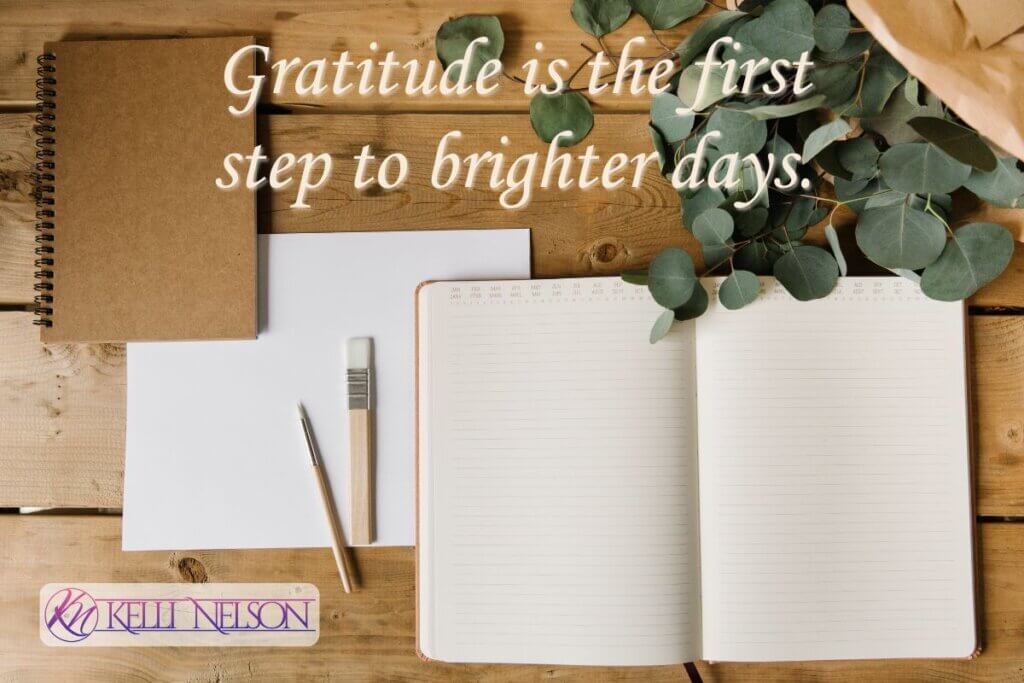 Gratitude An open notebook with blank pages, a pencil, a brush, and a closed journal on a wooden surface, with eucalyptus leaves nearby. The quote 'Gratitude is the first step to brighter days.' — Kelli Nelson is overlaid, and a logo is in the bottom left corner.