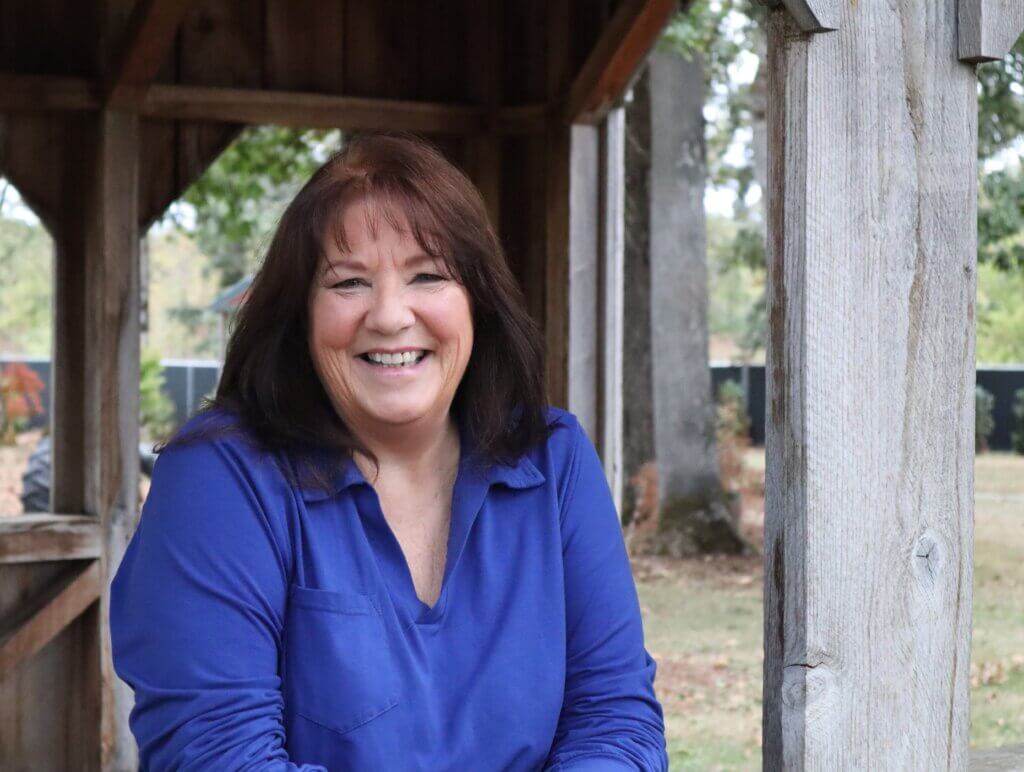 Kelli Nelson smiling while seated under a wooden outdoor structure, wearing a blue blouse.