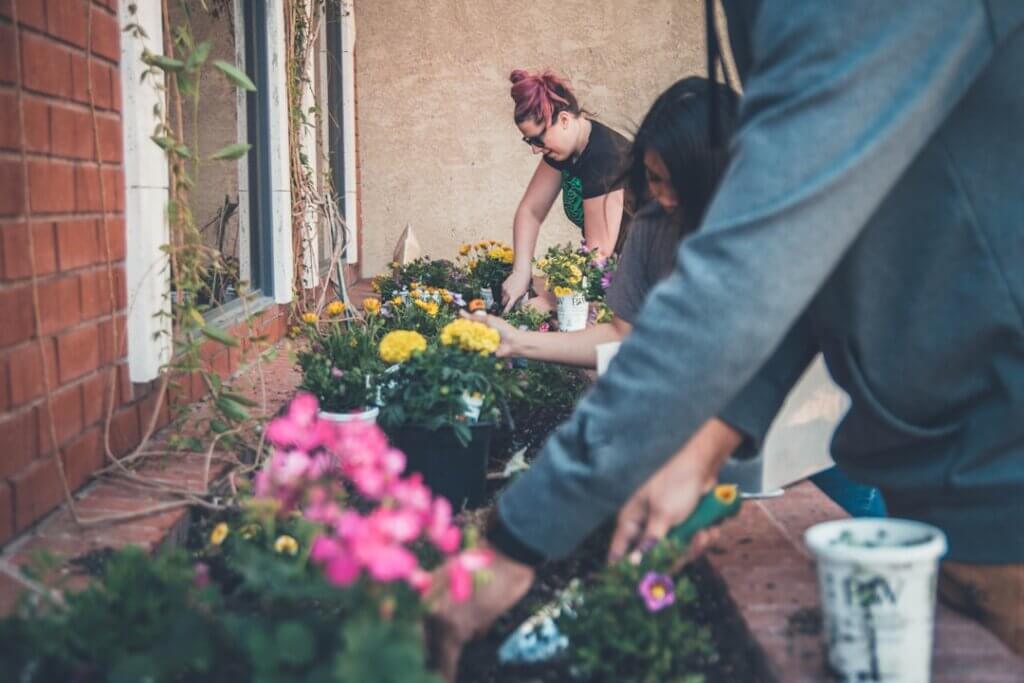 Three people planting flowers together outside a brick home, tending soil and working side by side.
