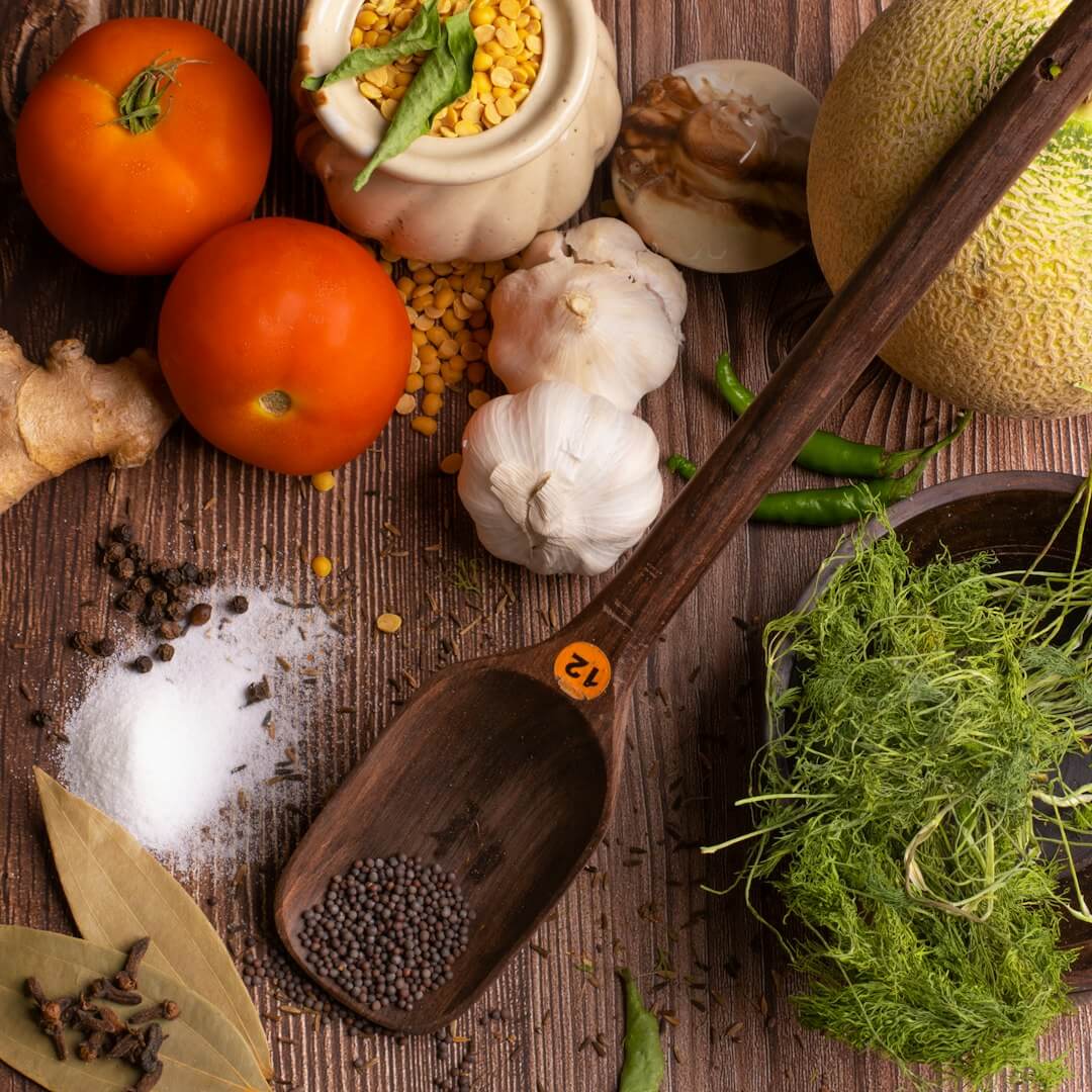Fresh vegetables, herbs, and spices, including tomatoes, garlic, ginger, and fennel, arranged on a wooden table with a wooden spoon